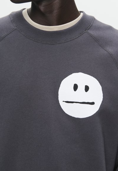 Grauer Pullover mit einem runden, weißen grafischen Gesichtsmuster, das zwei schwarze Punkte als Augen und eine gerade Linie als Mund zeigt.