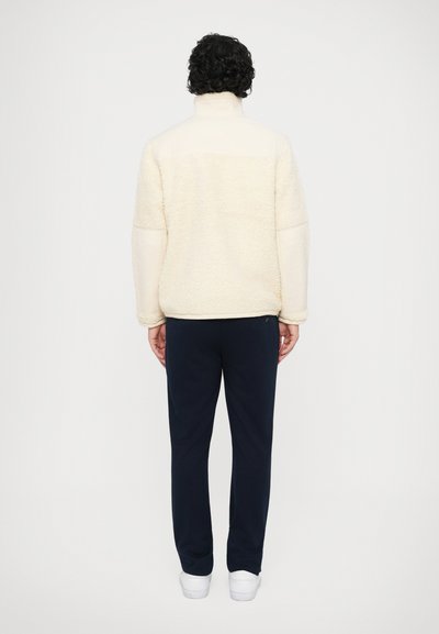 Pólo de polar em creme com gola alta e textura suave, combinado com calças de moletom azul-marinho e ténis brancos, visto pela parte de trás.