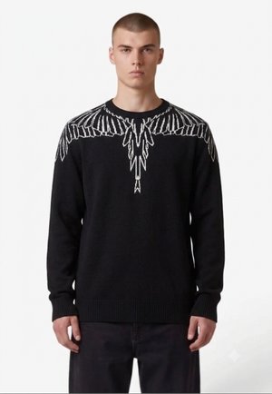 Giovane uomo che indossa un maglione nero con un motivo di ali bianche sulle spalle e sulla parte superiore del petto, in piedi davanti a uno sfondo bianco semplice.