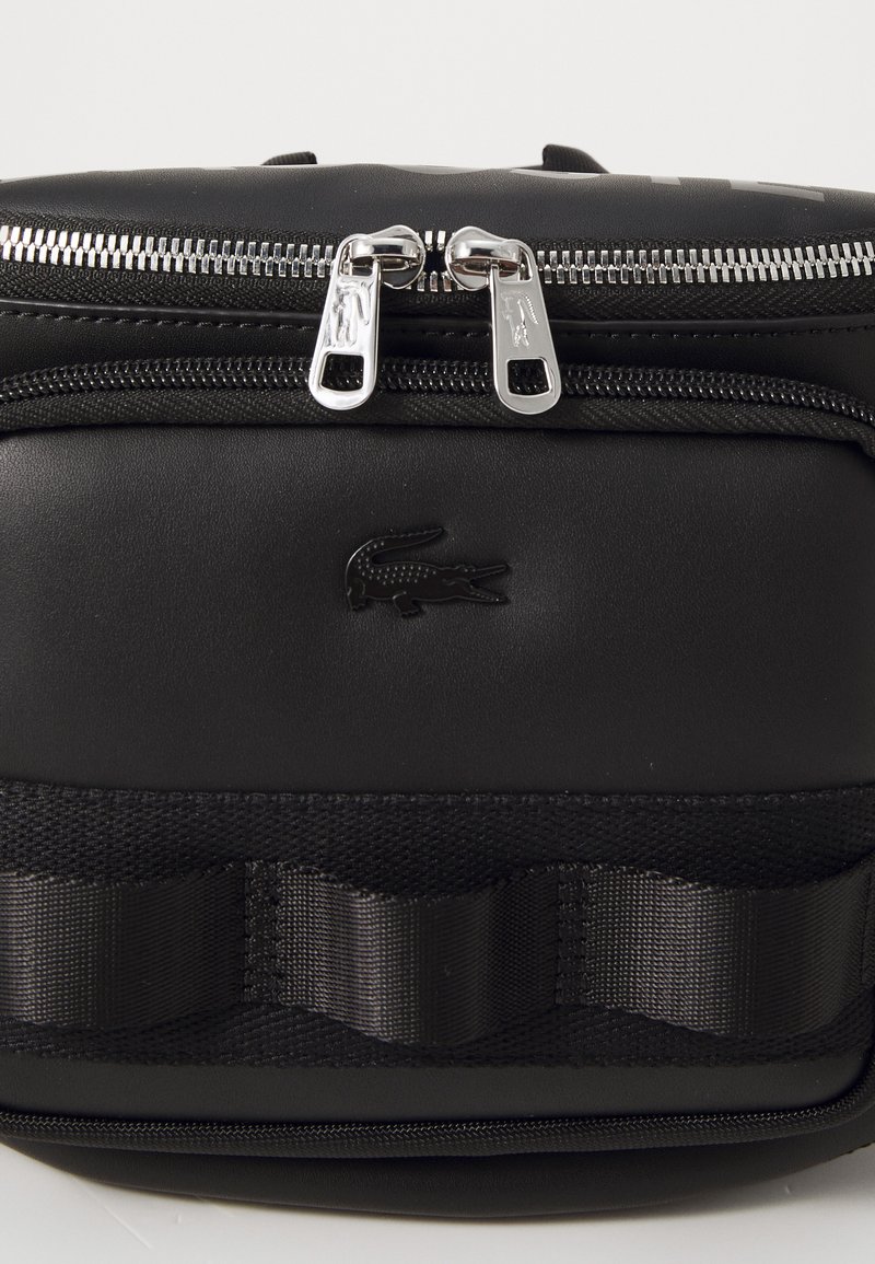 Sac synthétique noir avec deux zips argentés, une bandoulière texturée et un logo crocodile embossé à l'avant.