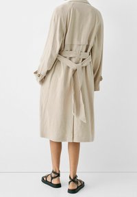Beżowy płaszcz z lnu z paskiem w talii, z długimi rękawami i luźnym krojem. Połączony z czarnymi sandałami na paski.