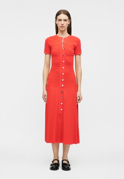 Γυναίκα που στέκεται όρθια φορώντας ένα φωτεινό κόκκινο, με κουμπιά, μίντι φόρεμα με κοντά μανίκια και μαύρα επίπεδα παπούτσια, σε απλό λευκό φόντο.