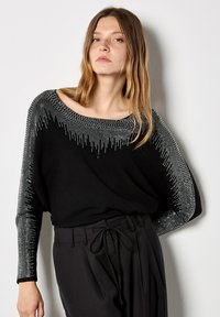 Giovane donna che indossa un maglione nero con motivi a gradiente di pois bianchi sulle maniche e sul colletto, abbinato a pantaloni neri a vita alta legati con un fiocco.