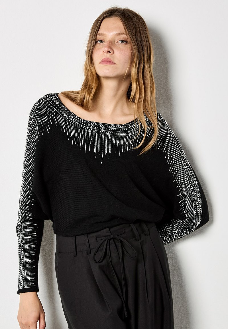 Giovane donna che indossa un maglione nero con motivi a gradiente di pois bianchi sulle maniche e sul colletto, abbinato a pantaloni neri a vita alta legati con un fiocco.