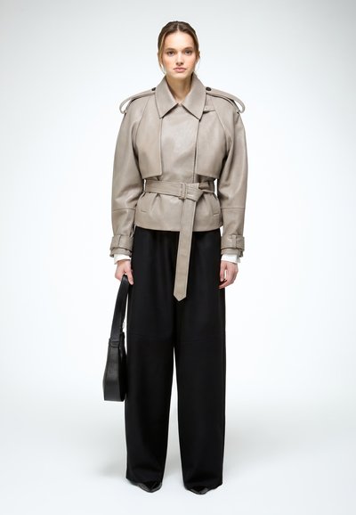 Nő, bézs öves bőrdzsekit és széles szárú fekete nadrágot visel, fekete táskát tart a kezében, tiszta fehér háttér előtt áll.