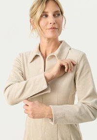 Rochie din catifea bej cu fermoar frontal, guler și mâneci lungi. Materialul prezintă o textură verticală, rigidsă și are o croială lejeră.