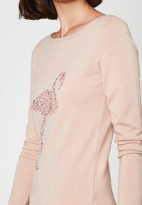 Kobieta w beżowej bluzce z długim rękawem, na której z przodu znajduje się różowa aplikacja z cekinami w kształcie flaminga.