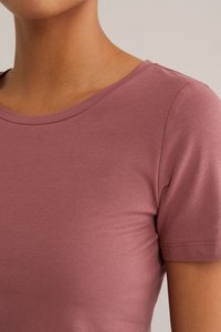 T-paita lyhyillä hihoilla, vaaleanpunaista väriä, valmistettu pehmeästä kankaasta. Sisältää pyöreän kaula-aukon ja sileän pinnan, jossa on minimaalista ommeltu yksityiskohtia.