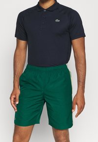 Marineblaues Poloshirt mit Kragen und drei Knöpfen, kombiniert mit grünen Sportshorts, die seitliche Streifen und einen elastischen Bund haben.