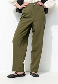 Pantalons vert olive taille haute avec une coupe droite et des plis à l'avant, associés à des chaussures plates noires avec des broderies florales.