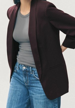 Personne portant une chemise grise ajustée, un blazer bordeaux à fines rayures et un jean bleu, une main posée sur la hanche.