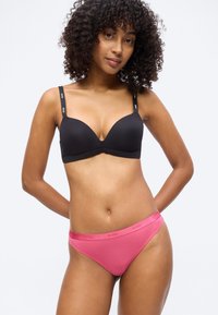 Γυναίκα με σγουρά μαλλιά που φοράει μαύρο σουτιέν με λουράκια με επωνυμία και ροζ εσώρουχο στυλ μπικίνι, στέκεται μπροστά σε λευκό φόντο.