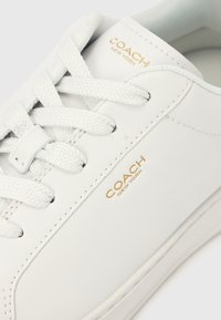 Pantof sport din piele albă cu șireturi texturate, având un logo auriu pe lateral și un design superior neted, evidențiind detalii minimaliste.