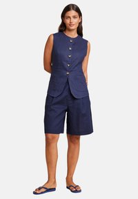 Navy linnevest med seks knapper, kombinert med matchende shorts til knelengden. Glatt tekstur, uten mønstre. Brukt med blå flip-flops.