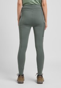 Groene strakke leggings van een gladde, rekbare stof met een zijzak, gecombineerd met bruine wandelschoenen met veters en stevige zolen.