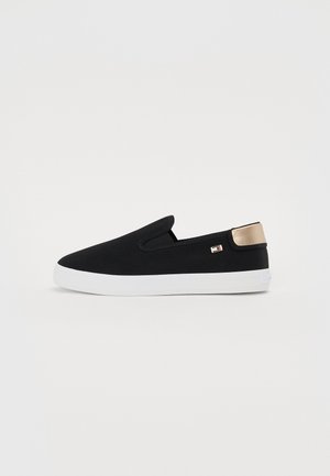 Czarny but typu slip-on z białą podeszwą i metalicznym złotym elementem na pięcie, z małym logo marki po boku.