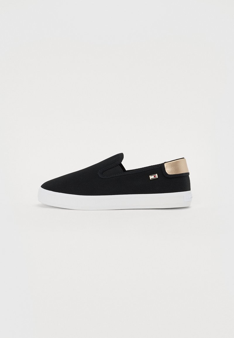 Sneakers slip-on noirs avec semelle blanche et languette de talon métallique dorée, avec un petit logo de marque sur le côté.