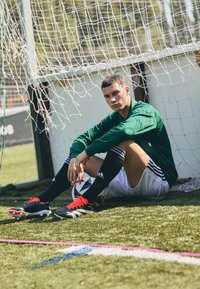 Nogometaš v zeleni majici z dolgimi rokavi, črnih kratkih hlačah in črnih nogavicah z belimi in rdečimi čevlji sedi na umetni travi blizu vrat.