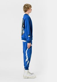 Sudadera azul con un gráfico de alas en la parte posterior, combinada con pantalones azules que tienen detalles de alas en blanco y azul. Botas blancas gruesas.