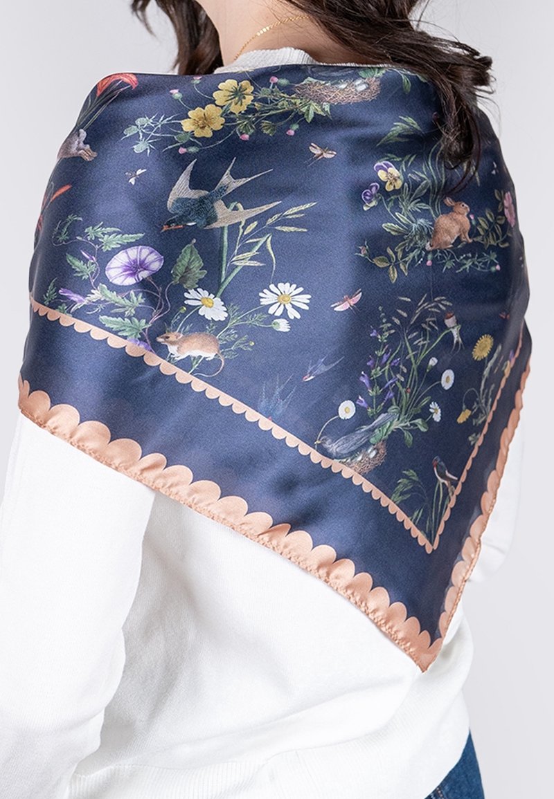 Femme portant un foulard en soie bleu marine avec des motifs floraux, oiseaux, souris et lapins et une bordure festonnée pêche sur un pull blanc.