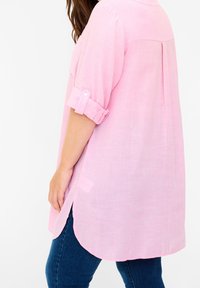 Blusa rosa chiaro a maniche corte con polsino abbottonato, orlo curvo e piega sul retro. Il tessuto sembra morbido e leggero.