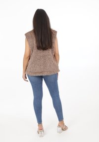 Bezrękawnik o puszystej strukturze w brązowym kolorze, zestawiony z dopasowanymi niebieskimi dżinsami oraz przezroczystymi sandałami na blokowym obcasie.