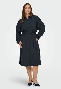 Ženska v obleki dolgem rukavu, do kolen, mornarsko modrih prugah, z pasom in temnimi visokimi petami, stoji ob enobarvni ozadju.