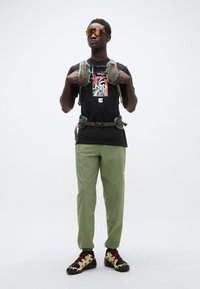 Μαύρο μπλουζάκι με γραφικό σχέδιο, πράσινο τακτικό σακίδιο, πράσινο εφαρμοστό παντελόνι και μαύρα παπούτσια με κίτρινες λεπτομέρειες και κόκκινα κορδόνια.