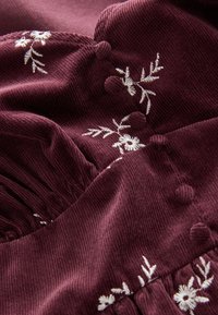 Tessuto in velluto a coste color bordeaux con motivi floreali bianchi ricamati e bottoni testurizzati, con dettagli plissettati lungo l'orlo.