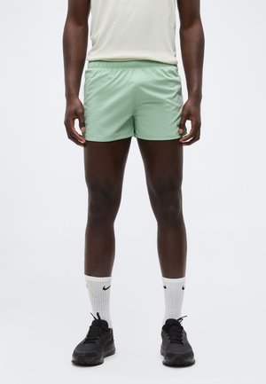 Mand iført lysegrønne træningsshorts, hvide Nike-strømper og sorte sneakers, stående mod en ensfarvet baggrund.