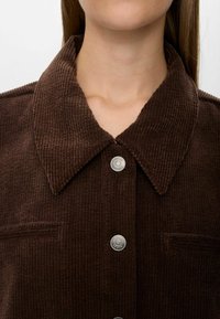 Close-up of brown corduroy jacket with large collar and silver snap buttons, worn by a person with long hair.