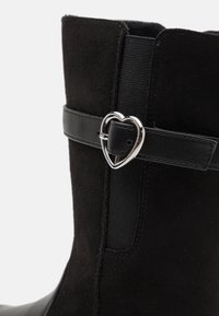 Botte en daim noir avec une sangle noire ornée d'une boucle en forme de cœur argenté sur le côté.