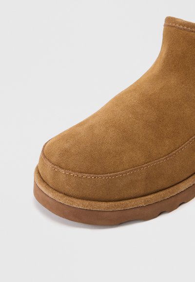 Botă ankle din suede maro, cu vârf rotunjit, detalii subtile de cusătură și talpă din cauciuc. Textura pare moale și netedă.