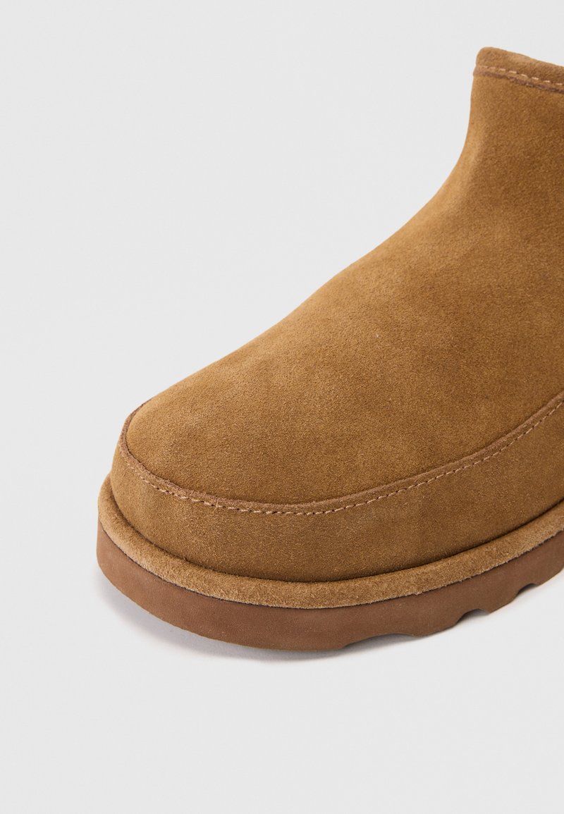 Bottine en suédine marron avec un bout rond, des détails de couture subtils et une semelle en caoutchouc. La texture semble douce et lisse.