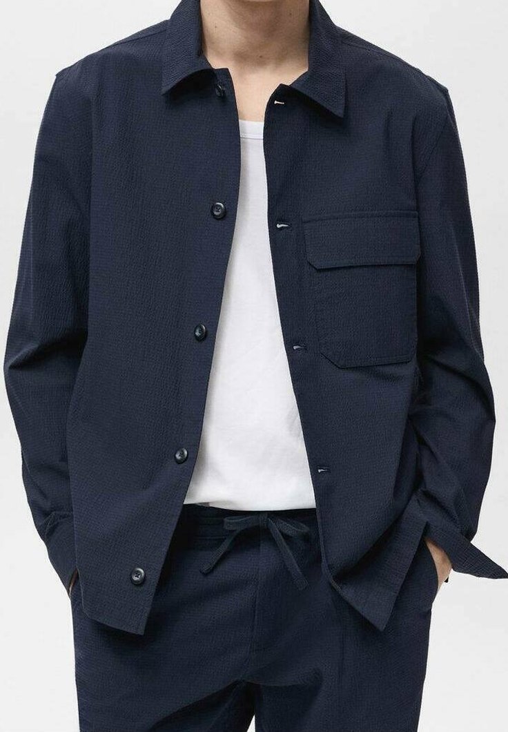 Navy blue button-up jacket featuring a textured fabric, a single pocket, and a relaxed fit. Worn over a white T-shirt with drawstring pants.