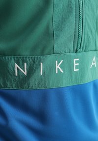 Nike-jacka i grön och blå med dragkedja framtill. Har ett mjukt, lättviktigt tyg med en framträdande vit logotyp på bröstet.