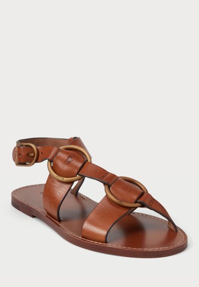 Brūna ādas sandale ar apaļām metāla detaļām, regulējams potītes siksniņa, atvērta pirksta dizains un plakana zole ar šuvēm.