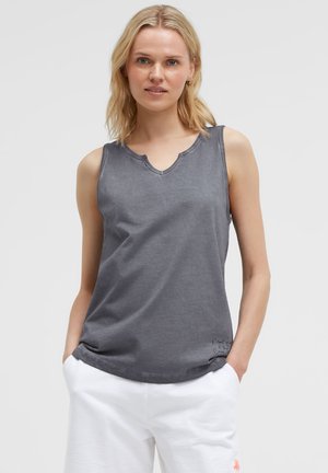 Blonde woman wearing a sleeveless gray top with a V-notch neckline and white pants, standing with hands in pockets against a plain background.