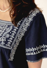 Haut bleu marine avec des broderies légères bleu clair le long de l'encolure et des manches, présentant des motifs floraux et à volutes sur un tissu doux.