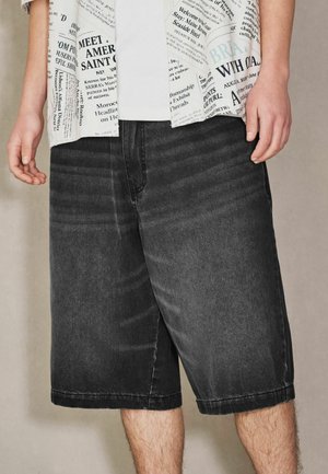 Mann trägt schwarze, knielange Denim-Shorts und ein weißes Hemd mit Zeitungsdruck, steht mit entspannten Händen seitlich.