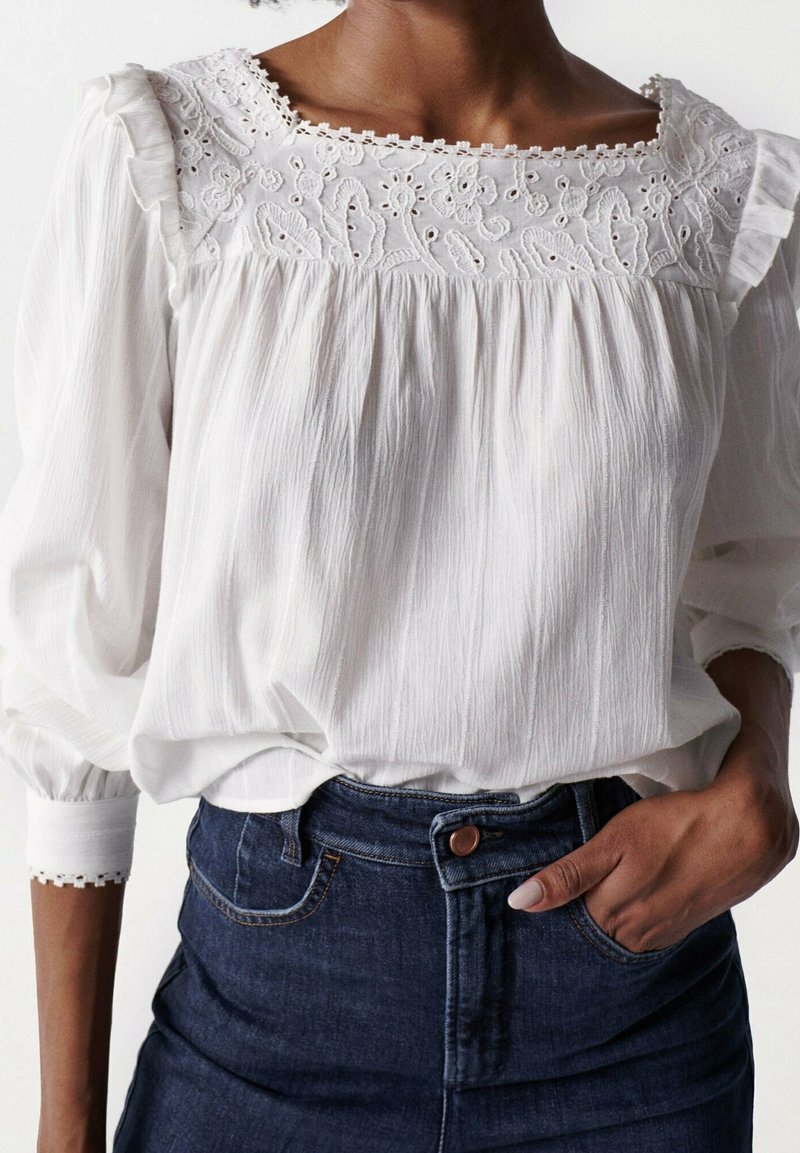 Weiße Bluse aus strukturiertem Stoff, mit Spitzenverzierungen am quadratischen Ausschnitt und Puffärmeln. Kombiniert mit einem dunklen Jeansrock.
