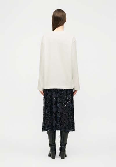 Γυναίκα με μακριά καστανά μαλλιά, ντυμένη με λευκό μακρυμάνικο τοπ, μαύρη φούστα μέσης με παγιέτες και μαύρες μπότες με τακούνι, στέκεται με την πλάτη προς τα εμπρός σε λευκό φόντο.