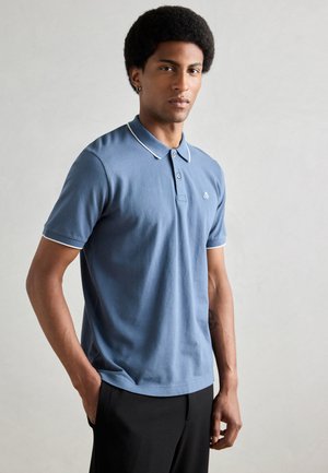 Camisa polo azul de manga corta, con cuello y acentos en las mangas en blanco, presentando un pequeño logo en el pecho. Combinada con pantalones negros.