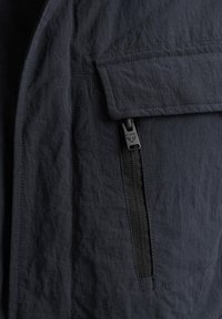 Marinejacke aus einem strukturierten, leichten Stoff mit einer Reißverschlusstasche und einer aufgesetzten Tasche, die klare Linien und subtile Nähdetails zeigt.