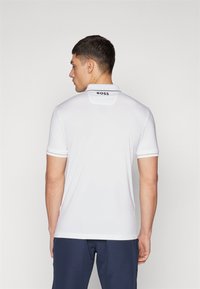 Biely polo tričko s krátkym rukávom a golierom, na zadnej strane s čiernym logom "BOSS" a kontrastnými pruhmi na rukávoch. Hladká textúra.