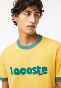 Żółta koszulka z turkusowymi wykończeniami, z wyraźnym napisem "Lacoste" oraz małym zielonym logo krokodyla. Wykonana z bawełny o gładkiej fakturze.