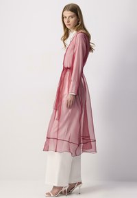Robe longue rose transparente avec une taille à nouer, un design superposé et des manches longues, associée à un pantalon large blanc et des sandales blanches à lanières.
