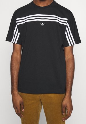 Person wearing a black Adidas t-shirt with white horizontal stripes across chest and sleeves, paired with brown pants.