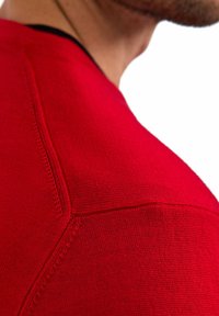 Vêtement en tricot rouge au design ajusté, présentant une texture lisse et des coutures détaillées le long de la couture des épaules.