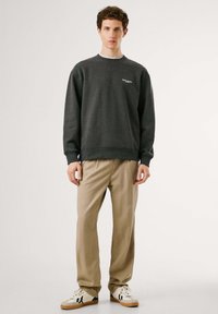 Sudadera gris con cuello redondo y detalle de logotipo, combinada con pantalones beiges. El modelo lleva zapatillas blancas con detalles en negro. Textura de tela suave.